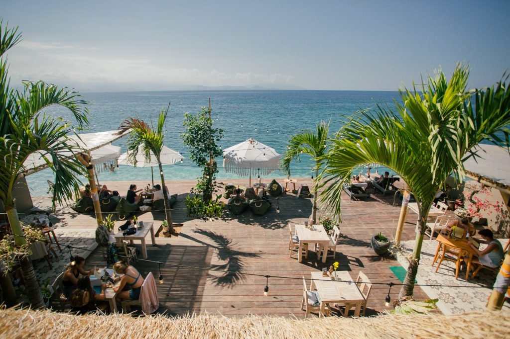 Dining Area deck at Penida Colada Beachbar overlooking the ocean, with palm trees, bean bags, umbrellas and beachfront views